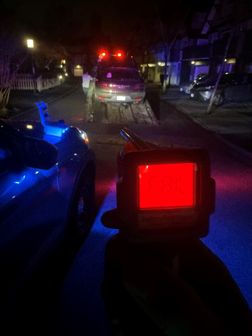 officer holding approved screening device showing a red light with the text: fail. A car on a flatbed tow truck is in the background.